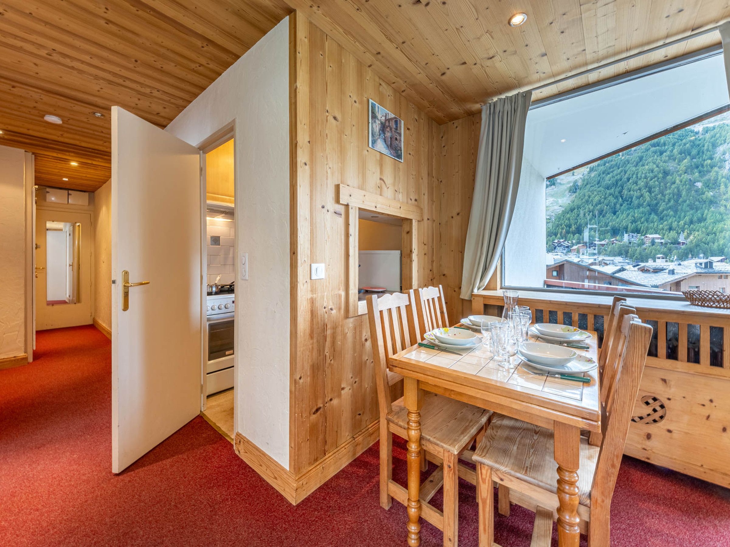 Un intérieur en bois avec une table à manger dressée pour quatre personnes, une cuisine visible à travers une porte ouverte et une fenêtre donnant sur une zone boisée.