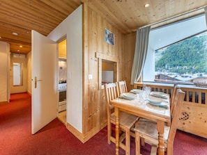 A wooden interior with a dining table set for four, a kitchen visible through an open door, and a window with a view of a forested area.
