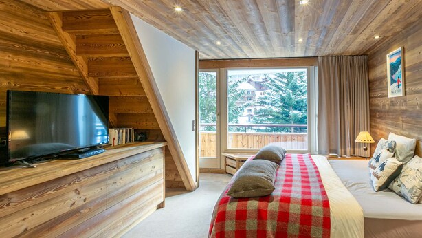A cozy bedroom with a wooden ceiling, a large window, a bed with a red and white checkered blanket, and a flat-screen TV on a wooden dresser. - Spacious 3-BR Duplex with South Balcony, WiFi, Parking, Ski Storage, Near Slopes (Val-d'Isère)