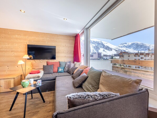 A modern living room with a large window, wooden walls, a sofa with multiple cushions, a small table with a lamp, and a flat-screen TV mounted on the wall.