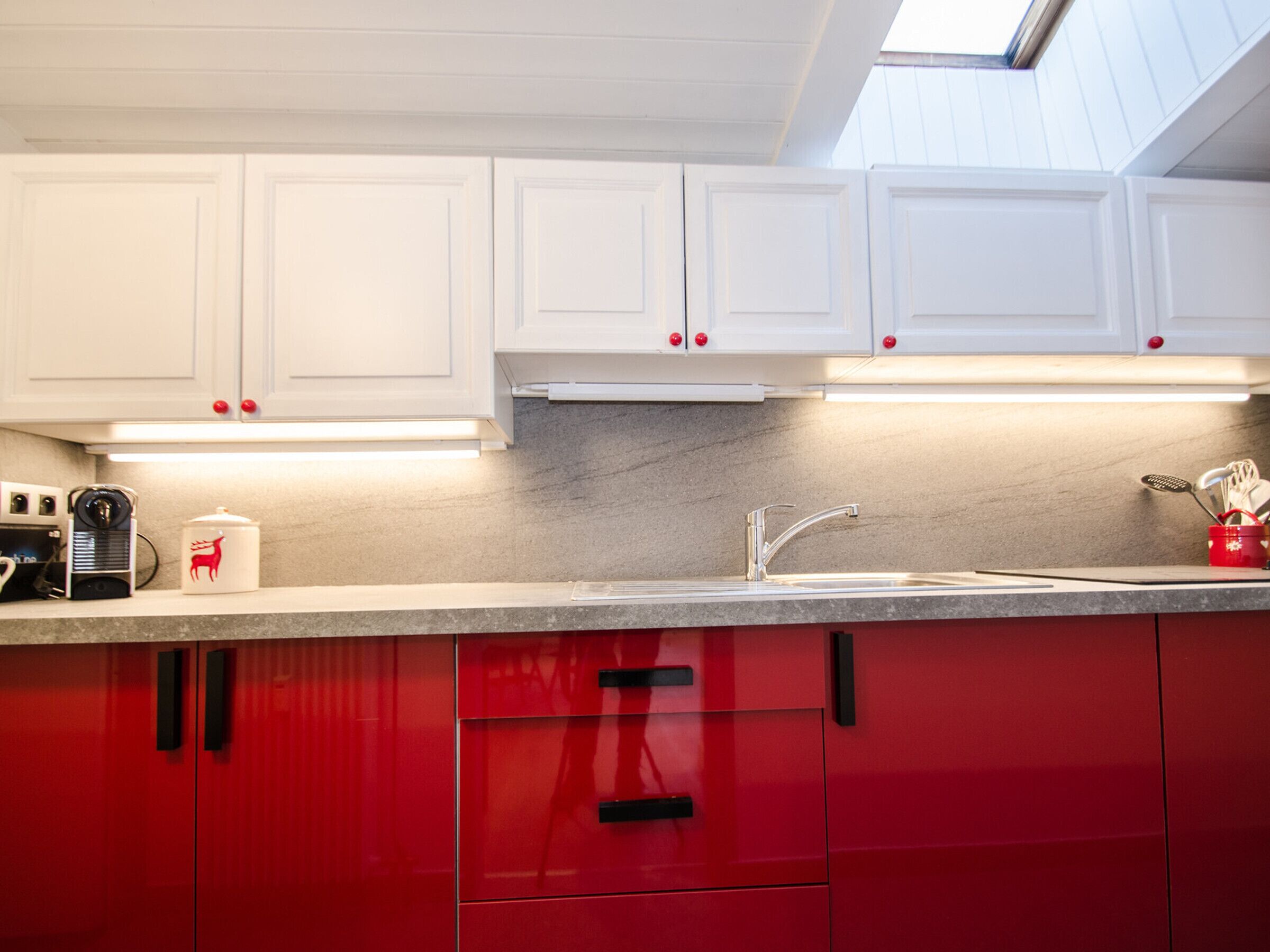 A kitchen with white upper cabinets, a red lower cabinet, a stainless steel sink, and a skylight above.