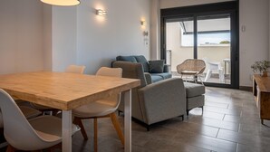 Un salon moderne comprenant un coin repas, un ensemble de canapés, un fauteuil en osier et une vue à travers une porte vitrée.