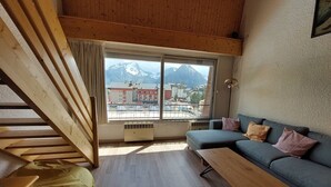 Una habitación con techo de madera, un ventanal con vistas a la montaña, un sofá azul, una mesa de madera y una escalera.