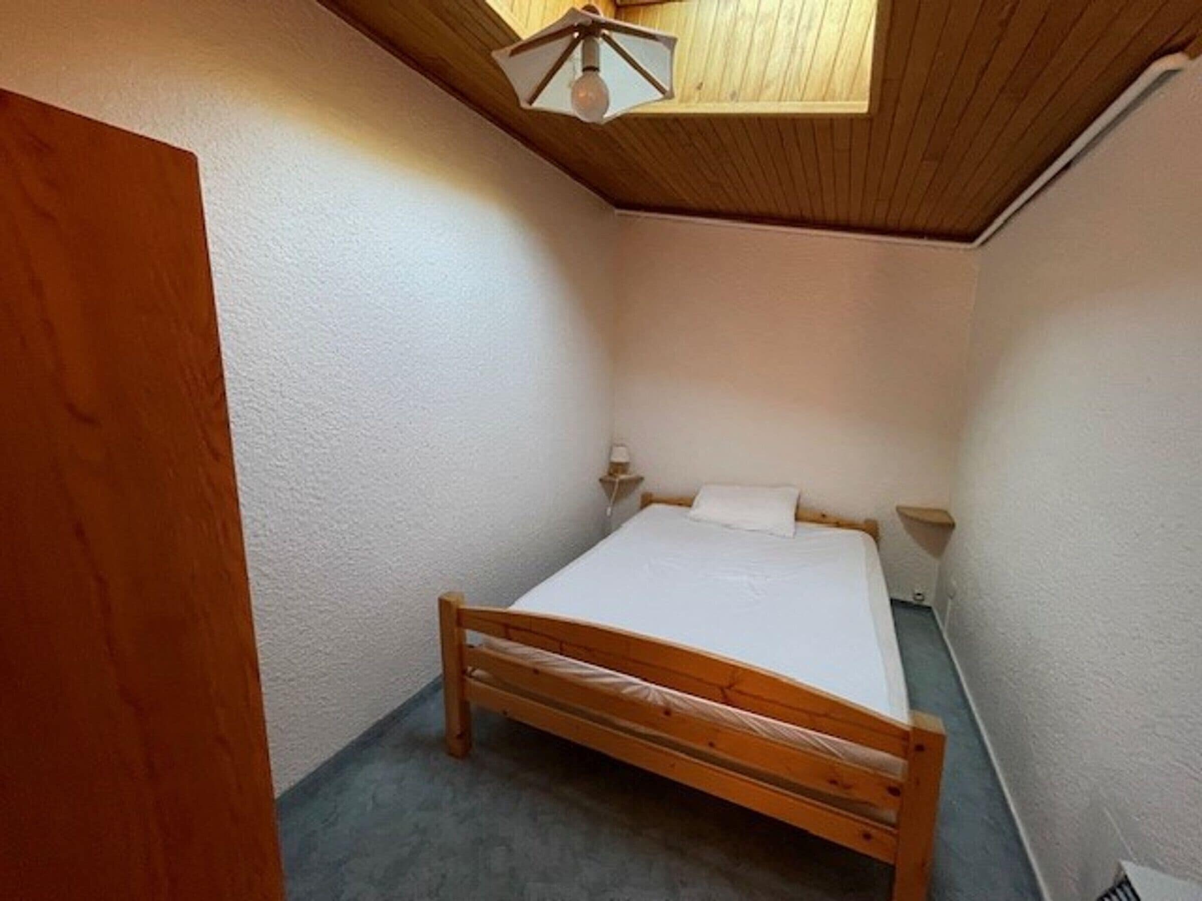 Una habitación pequeña con cama individual, techo y paredes de madera, una mesita de noche y una mesilla.