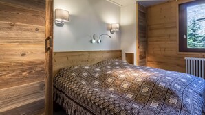 Une chambre dans une cabane en bois, avec une couvre-lit à motifs, deux lampes fixées au mur et une fenêtre donnant sur un paysage verdoyant.