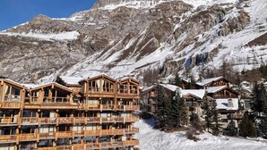 Un hôtel de style chalet en bois, doté de nombreux balcons, qui se dresse sur fond de montagnes enneigées et d’un ciel d’un bleu éclatant.