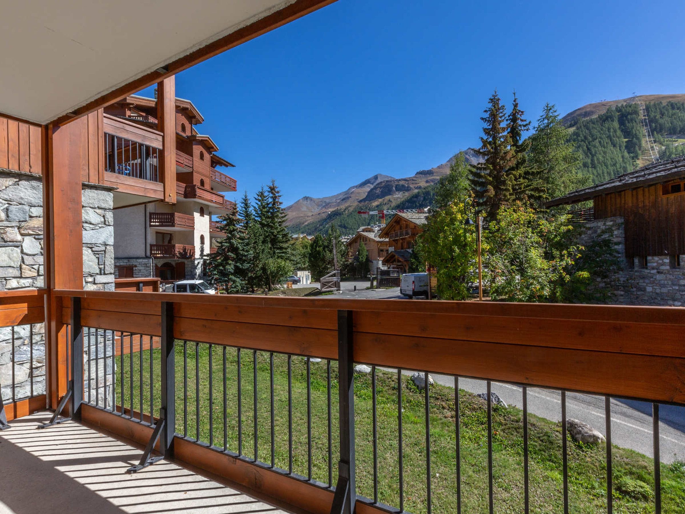 A wooden balcony with a stone wall, overlooking a mountainous landscape with buildings and trees.