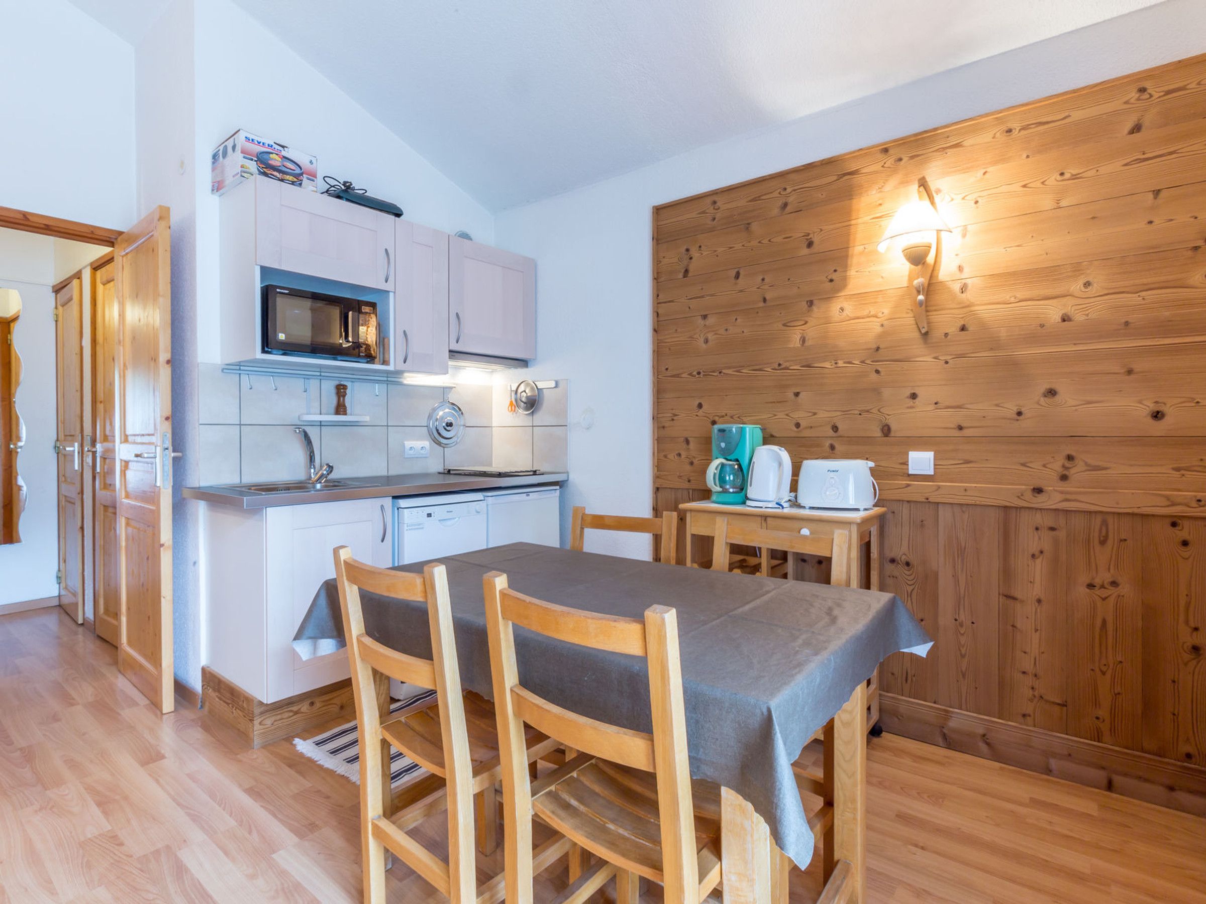 A compact kitchen with a microwave, toaster, and coffee maker, a dining table with chairs, and a wooden wall with a light fixture.