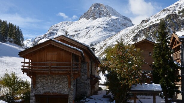 Un chalet de montagne à l’architecture en pierre et en bois, situé au pied de sommets enneigés, avec un ciel dégagé en toile de fond.