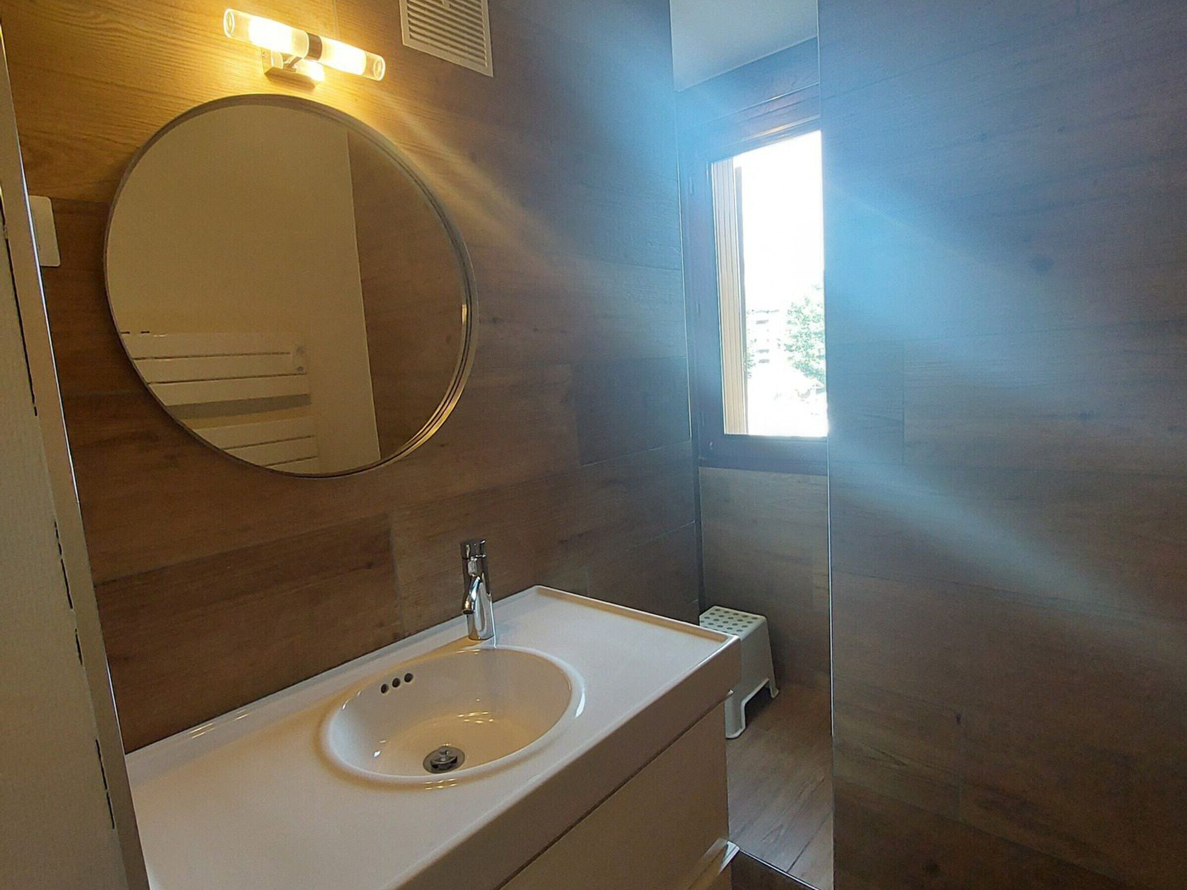 A bathroom with a round mirror, a white sink, and a window.