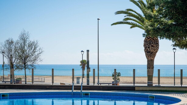 A swimming pool with a wooden deck, a palm tree, and a view of the beach and ocean. - Seaside apartment with terrace and communal pool (Torredembarra)