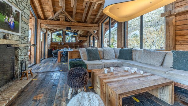 A rustic cabin interior with a wooden ceiling, stone walls, a large window, a sofa, a coffee table, and a dog.