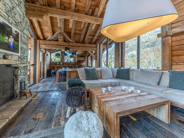 A rustic cabin interior with a wooden ceiling, stone walls, a large window, a sofa, a coffee table, and a dog.