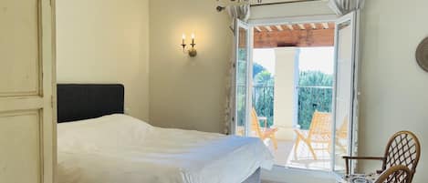 A bedroom with a bed, a chandelier, a balcony with chairs, and a view of greenery.
