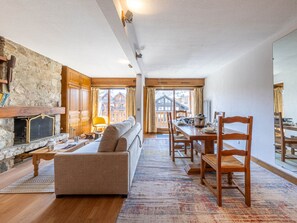 Un salón acogedor con chimenea de piedra, muebles de madera y vistas a un albergue de montaña a través de amplias ventanas.
