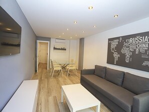 Una sala de estar moderna con un sofá gris, una mesa de centro blanca y una mesa de comedor de vidrio con sillas. Se observa una obra de arte en la pared que representa un mapa del mundo y un televisor de pantalla plana montado en la pared.