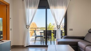 Una sala de estar moderna con un sofá, una mesa de centro y un televisor sobre un mueble bajo. Se ve un balcón a través de una puerta corrediza de vidrio con cortinas.