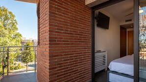 Un balcón con pared de ladrillo, una cama con ropa blanca, un televisor montado en la pared y vistas a árboles y edificios.