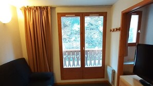 Una stanza con un divano scuro, una scrivania in legno con un televisore, e una porta che dà su un balcone esterno con ringhiera.