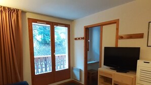 Un soggiorno con tappeto blu, una TV su un supporto in legno, una porta scorrevole affacciata sul verde e una tenda marrone.