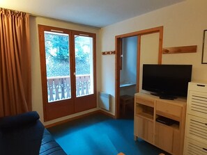A living room with a blue carpet, a TV on a wooden stand, a sliding door with a view of greenery, and a brown curtain. - Central studio with cabin for 4, balcony (Valfréjus)