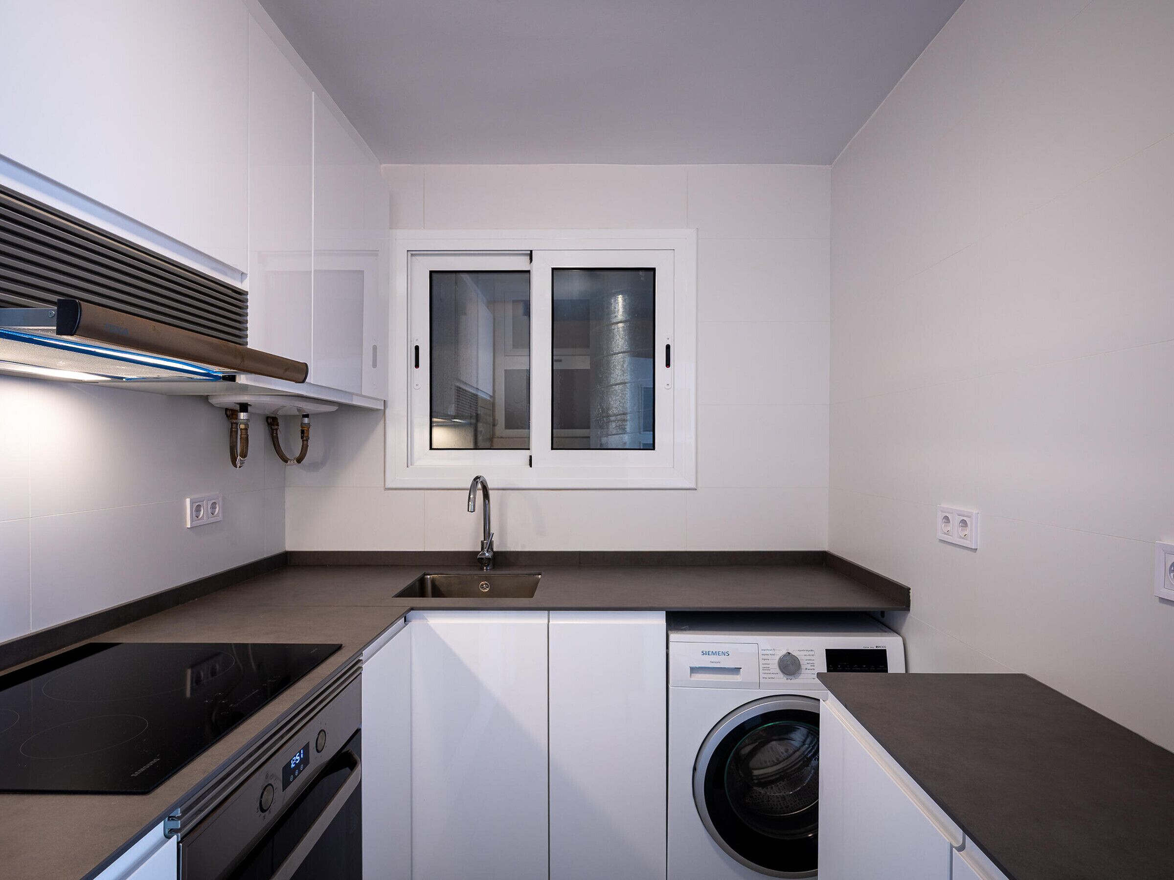 Cocina compacta con lavadora y secadora integradas, una ventana y un lavamanos de acero inoxidable.