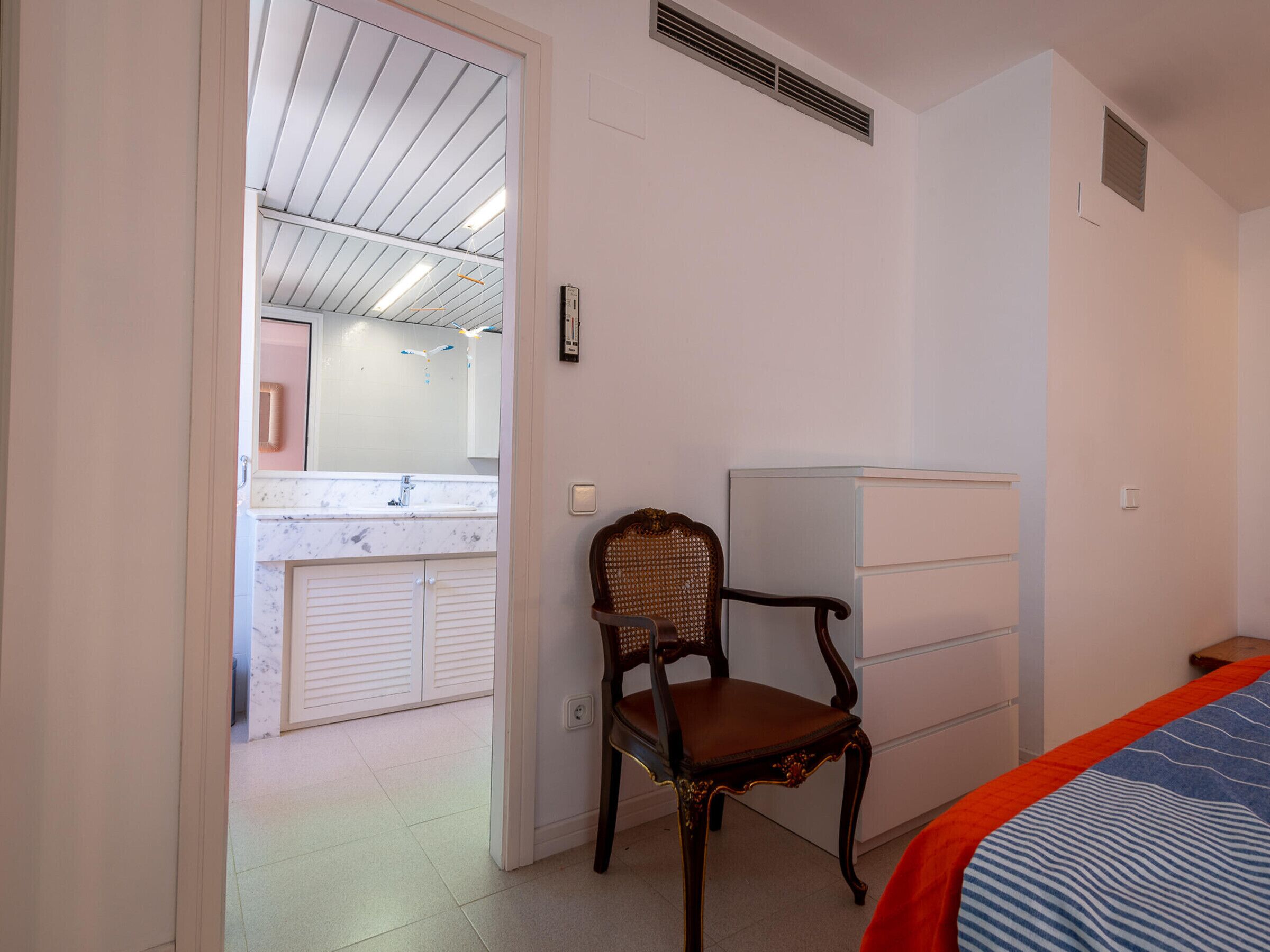 A bedroom with a bed, a chair, a wardrobe, and a bathroom visible through an open door.