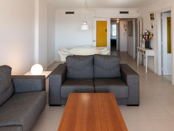 A modern living room with a grey sofa, a wooden coffee table, and a white dining table.