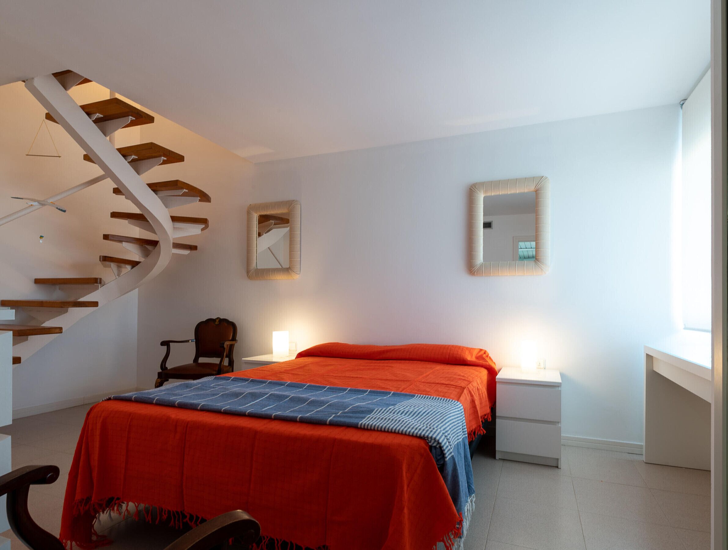 Una camera da letto con una scala a chiocciola, un letto con copripiumino arancione, una sedia, un comodino con una lampada e uno specchio a parete.