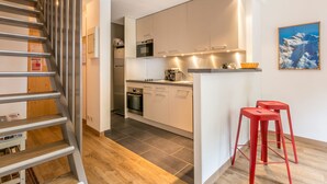 A modern kitchen with white cabinets, a stainless steel oven, and a microwave. There is a wooden staircase on the left and a red stool on the right.