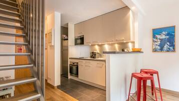 A modern kitchen with white cabinets, a stainless steel oven, and a microwave. There is a wooden staircase on the left and a red stool on the right.
