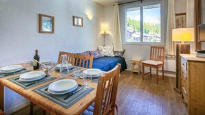 Ein Essbereich mit einem Holztisch, gedeckt für vier Personen, einem Bett mit blauer Bettdecke und einem Fenster mit Blick auf eine Berglandschaft.