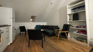 A compact living space with a wooden floor, a black sofa, a dining table with chairs, a white radiator, and a bookshelf with various items.
