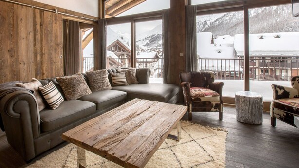 Una habitación acogedora con un sofá modular, una mesa de centro de madera y un sillón con una manta de piel. Amplias ventanas ofrecen vistas a un paisaje montañoso nevado.