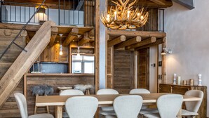 Un comedor rústico con una mesa de madera larga, seis sillas tapizadas, una araña con cuernos de ciervo y una escalera con barandillas de madera.