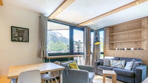 A modern living room with a wooden floor, a sofa, a small table, and a window with a view of mountains. - Elegant 3-Room Apartment with Balcony in Central Val-d'Isère – Sleeps 6, Modern Amenities (Val-d'Isère)