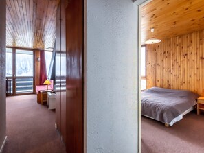 A hotel room with a wooden ceiling, a bed with a grey blanket, a bedside table with a lamp, and a view of a snowy landscape through a large window.