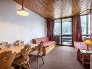 A cozy cabin interior with a wooden dining table, chairs, a sofa, and a window with a view of a snowy landscape.