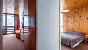 A hotel room with a wooden ceiling, a bed with a grey blanket, a bedside table with a lamp, and a view of a snowy landscape through a large window. - Charming 2-room apartment with pets allowed (Val-d'Isère)