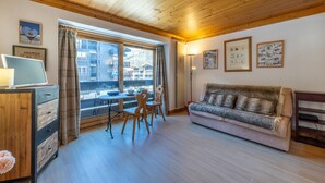A cozy living room with a wooden ceiling, a sofa, a dining table with chairs, and a view of buildings through large windows. - Cozy Studio with Wifi in Central Location, Close to Slopes, Pets Welcome (Val-d'Isère)