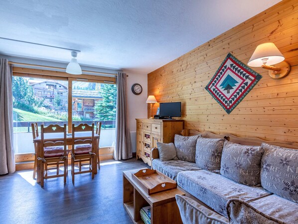 A cozy room with a wooden floor, a sofa with patterned cushions, a wooden cabinet, a television, and a large window offering a view of a building and trees. - Cozy, Well-Maintained Apartment at the Foot of the Slopes with Parking (Val-d'Isère)