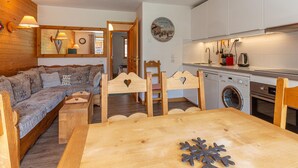A cozy living room with a sofa, wooden chairs, a dining table, and a kitchen area with a washing machine.