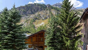 A wooden cabin with a balcony, surrounded by evergreen trees, set against a mountainous backdrop.