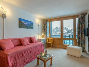 Um quarto aconchegante com um sofá vermelho, uma pequena mesa com uma xícara de café, um quadro de montanhas e grandes janelas com vista para montanhas nevadas.