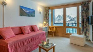 Ein gemütliches Zimmer mit einem roten Sofa, einem kleinen Tisch mit einer Kaffeetasse, einem Bergpanorama und großen Fenstern mit Blick auf schneebedeckte Berge.