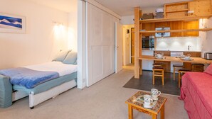 Ein kompakter Raum mit einem Bett, einem kleinen Tisch mit zwei Tassen, einer Küchenzeile und einer Tür zu einem weiteren Zimmer.