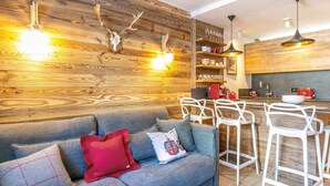 A modern kitchen with wooden accents, a grey sofa with red and white cushions, and white bar stools.