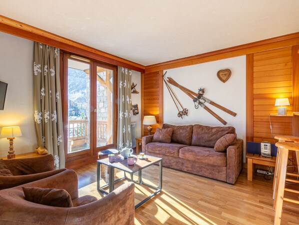 A cozy living room with a wooden floor, a sofa, a coffee table, and a fireplace. There are curtains on the windows, and a ski rack on the wall. - Charming 2-room apartment with WIFI in the heart of Val-d'Isère (Val-d'Isère)