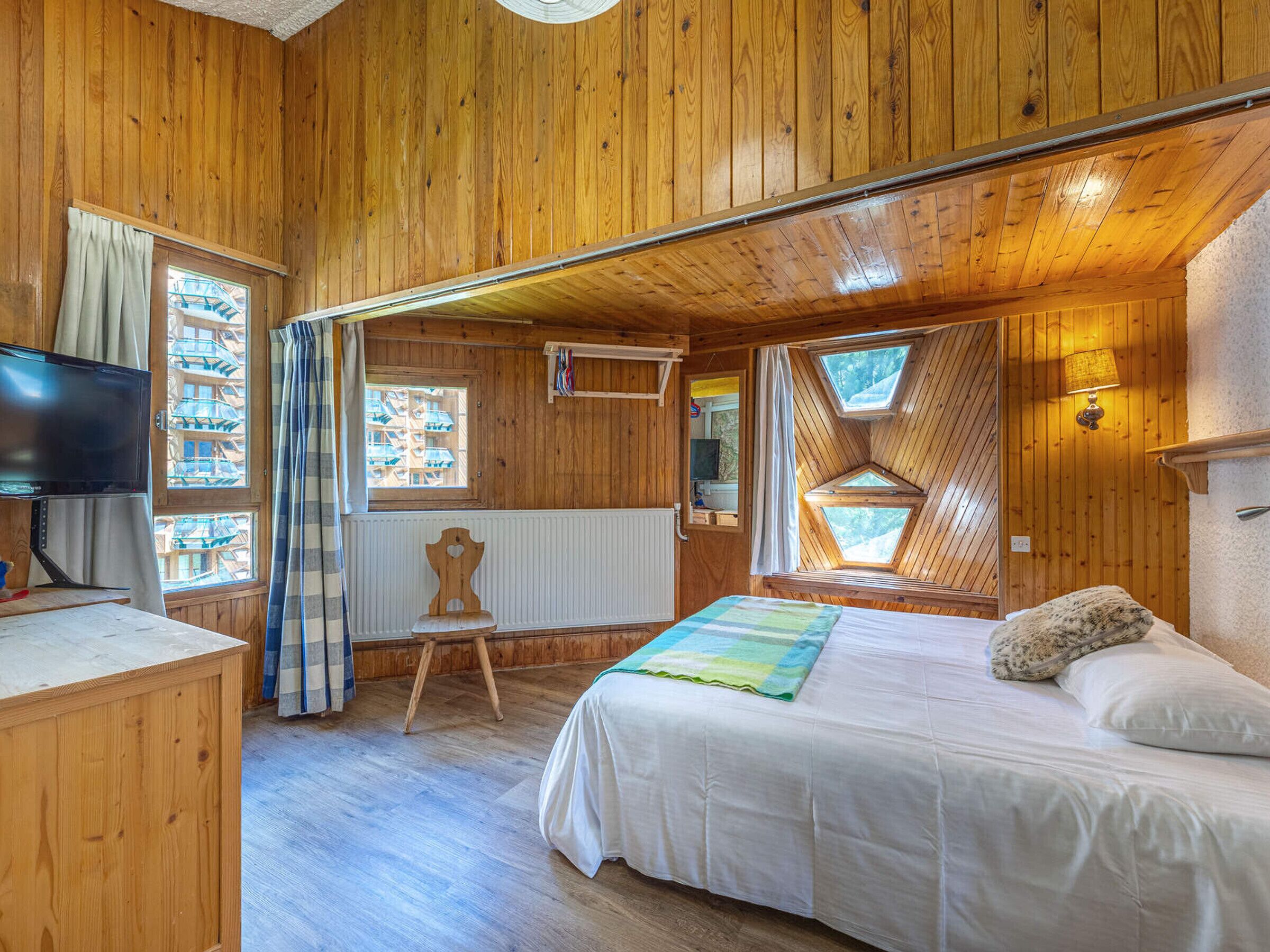 A wooden cabin bedroom with a bed, a chair, a television, and windows.
