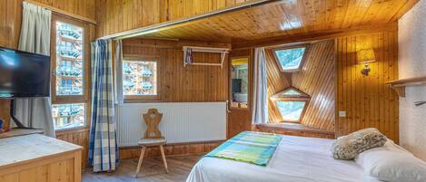 A wooden cabin bedroom with a bed, a chair, a television, and windows.